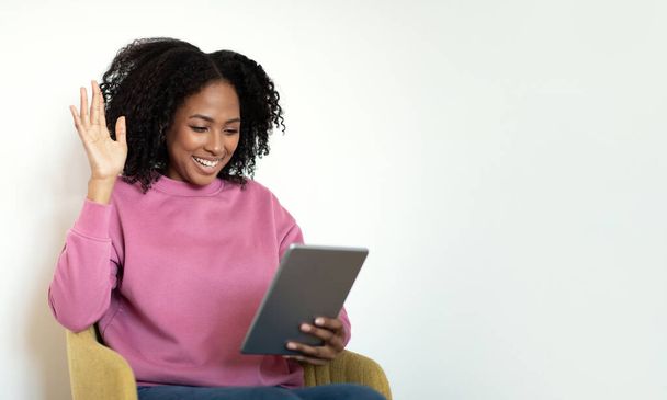 Cheerful millennial african american curly lady in casual sits on armchair, waving hand in tablet on white wall background. Greeting, hi, work and study, video call at home, ad, offer - Foto, imagen