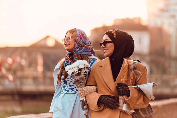 Pareja mujer una vistiendo un hijab y un vestido moderno pero tradicional, y la otra en un vestido azul y bufanda, caminando juntos por la ciudad al atardecer. Uno lleva un ramo y pan, mientras que el - Foto, imagen