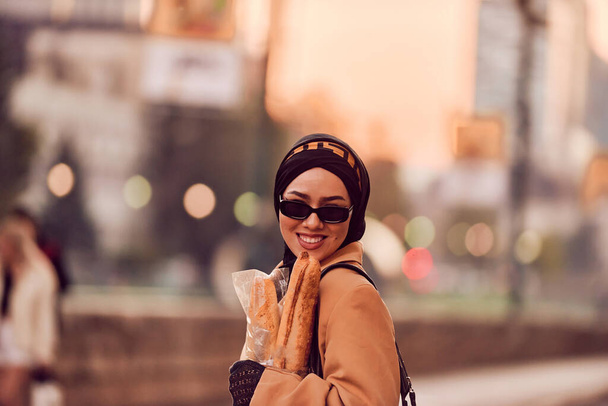 Una mujer hiyab, vestida con un lujoso atuendo vintage francés, mientras camina por las calles de la ciudad al atardecer, llevando pan, un ramo de flores y periódicos en su mano, evocando una cautivadora mezcla - Foto, imagen