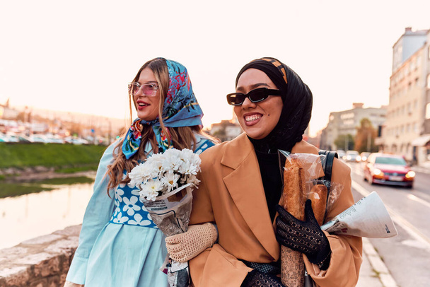 Пара женщин одета в хиджаб и современное, но традиционное платье, а другая в синем платье и шарфе, прогуливаясь вместе по городу на закате. Один несет букет и хлеб, в то время как - Фото, изображение