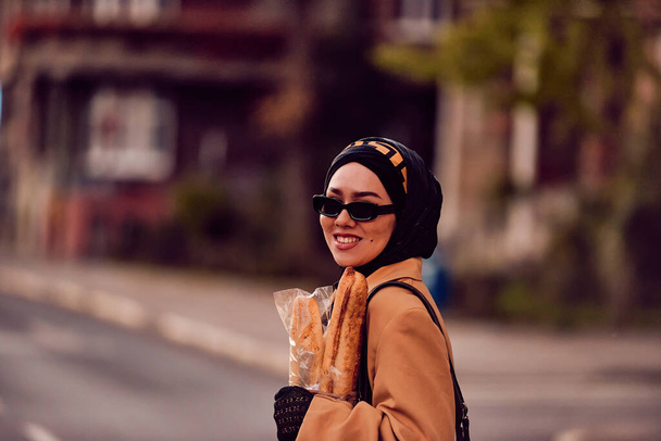 A hijab woman, donning a luxurious vintage French outfit, as she walks through the city streets at sunset, carrying bread, a bouquet of flowers, and newspapers in her hand, evoking a captivating blend - Foto, immagini