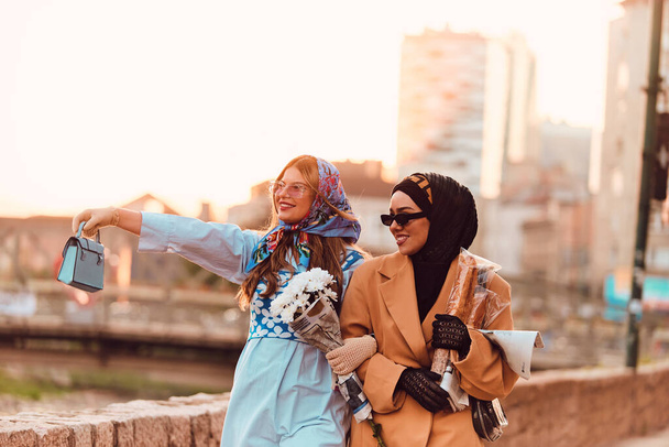 Eine Frau trägt einen Hijab und ein modernes, aber traditionelles Kleid, die andere ein blaues Kleid und einen Schal und läuft bei Sonnenuntergang gemeinsam durch die Stadt. Einer trägt einen Strauß und Brot, während der andere - Foto, Bild