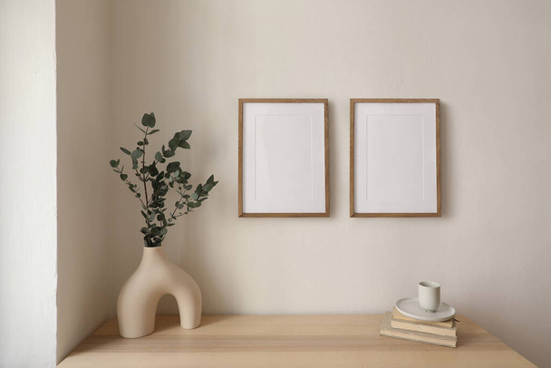 Two vertical picture frame mockup hanging on beige wall. Cup of coffee, books on wooden table. Vase with silver eucalyptus tree branches. Elegant interior, beautiful home decor, trendy working space. - Fotoğraf, Görsel