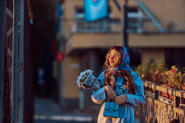 Fascino di una donna ornata in un abito tradizionale blu squisito, portando una borsa blu e un mazzo di fiori, passeggiando con grazia per la città al tramonto, creando una scena ipnotizzante di - Foto, immagini