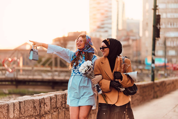 Couple woman one wearing a hijab and a modern yet traditional dress, and the other in a blue dress and scarf, walking together through the city at sunset. One carries a bouquet and bread, while the - Photo, Image