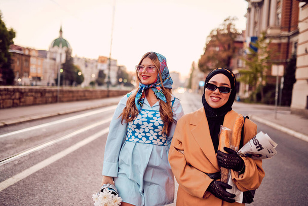 Pár žen v hidžábu a moderních, ale tradičních šatech, a druhá v modrých šatech a šále, procházející se společně městem při západu slunce. Jeden nese kytici a chléb, zatímco - Fotografie, Obrázek