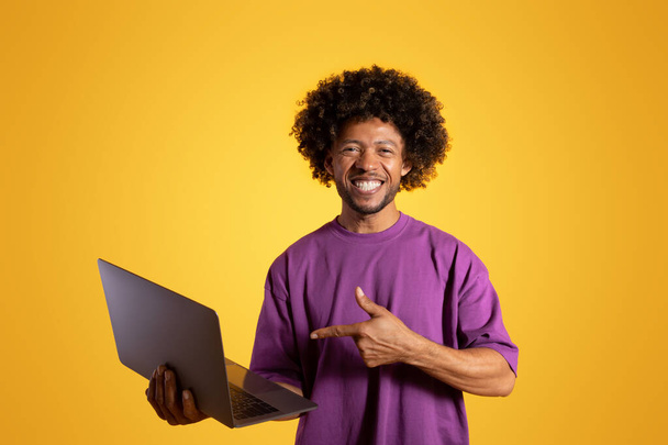 Cheerful black adult curly guy in purple t-shirt points finger at laptop, isolated on orange background, studio. Recommendation great news for work and study, advice, ad and offer - Photo, Image