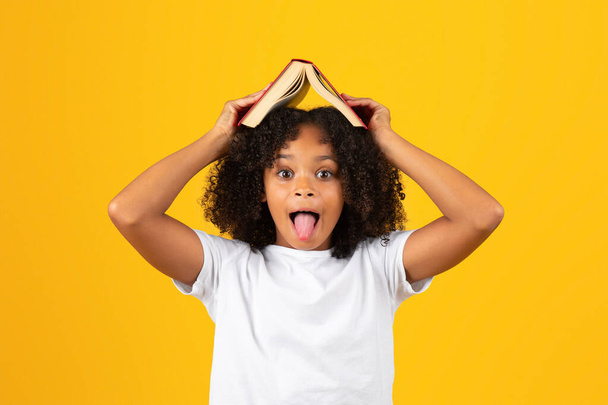 Portré vidám göndör tinédzser fekete iskolás lány fehér póló show nyelv, szórakoztató, tartsa könyv feje fölött, elszigetelt sárga háttér, stúdió. Tanulmány, tudás hobbi szabadidőben - Fotó, kép