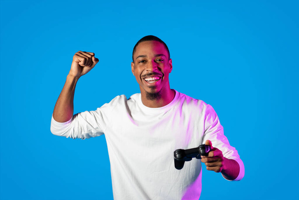 Emotional happy young handsome black guy wearing white shirt playing video game on blue background in neon light, champion holding joystick and clenching fist, copy space - Photo, Image
