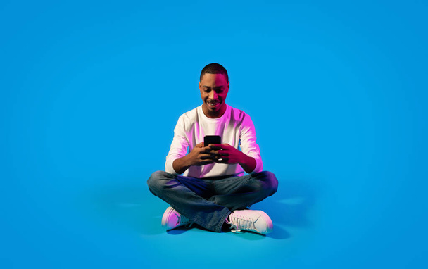 Cheerful happy young black guy in casual using modern cell phone and smiling over blue background in neon light, chatting with ladies on dating app, surfing on Internet. Gadget addiction concept - Zdjęcie, obraz