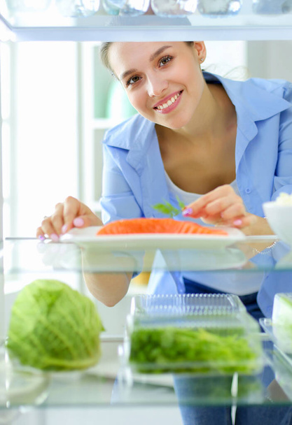Porträt einer Frau, die neben einem offenen Kühlschrank voller gesunder Lebensmittel, Gemüse und Obst steht. - Foto, Bild