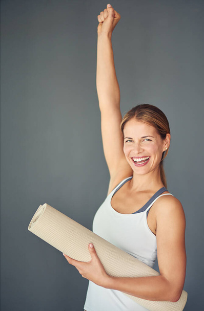 Happy woman, portrait and yoga mat with fist in celebration for healthy exercise against a grey studio background. Excited female person or yogi with smile for winning, fitness or workout success. - Photo, image