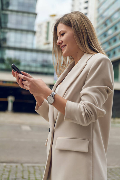 Side view of young female in elegant suit standing on city street and messaging on mobile phone - Foto, imagen