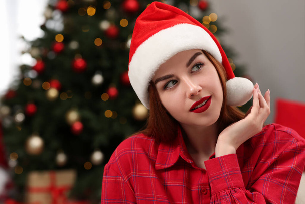 Beautiful young woman in Santa hat near Christmas tree indoors, space for text - Zdjęcie, obraz
