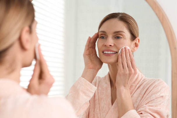 Beautiful woman removing makeup with cotton pad near mirror indoors - Fotoğraf, Görsel
