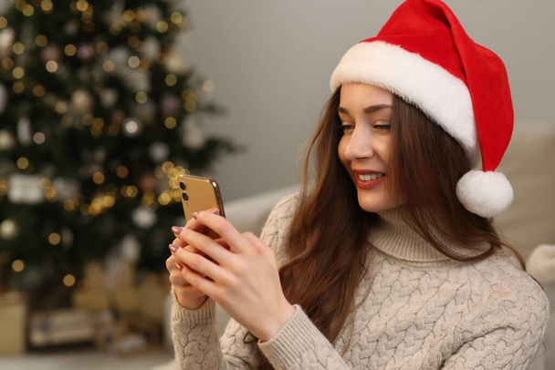 Beautiful young smiling woman in Santa hat using smartphone near Christmas tree at home, space for text - Photo, image