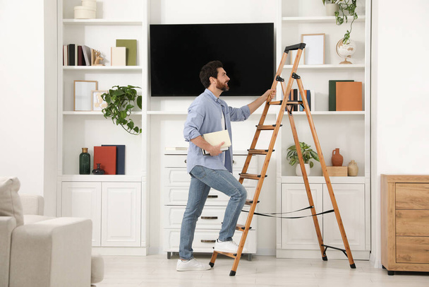 Man with books climbing on wooden folding ladder at home - Fotografie, Obrázek
