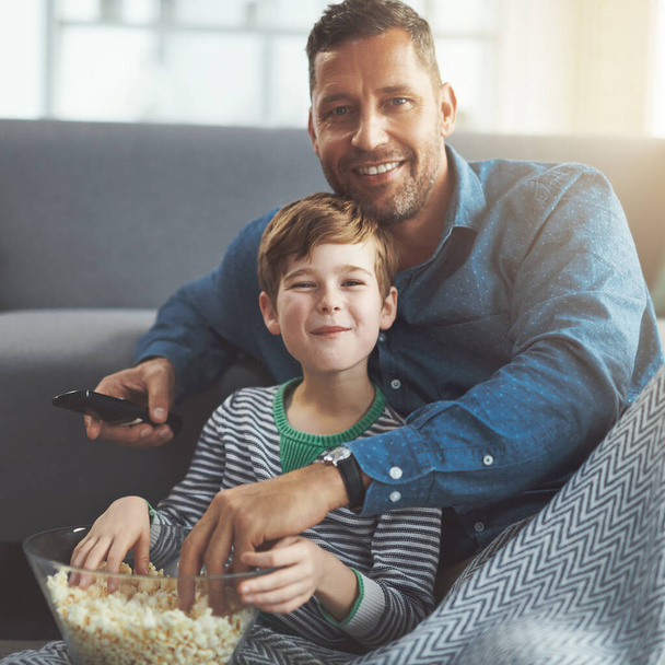 映画のポップコーンもある。床に座り、日中に自宅でポップコーンを食べながら一緒に映画を見ている屈託のない若い男の子と彼の父親の肖像画 - 写真・画像