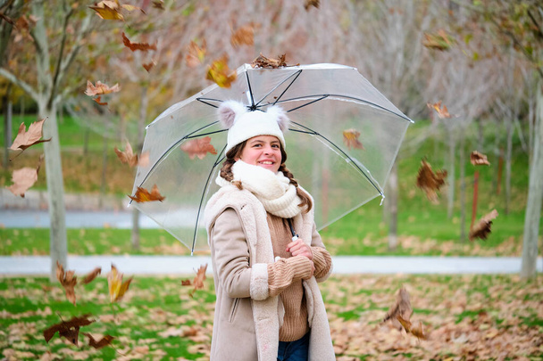 Preteen girl with an umbrella smiling and looking at camera while leaves are falling outdoors in autumn. - Photo, image