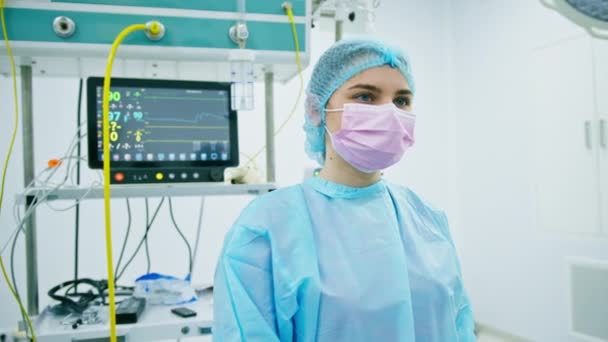 手術後の手術室に立つマスクと滅菌防護服を着た若い女性医師の肖像 - 映像、動画