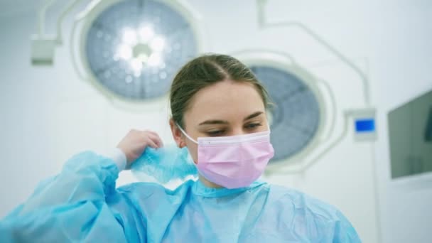 Retrato de una joven doctora con ropa protectora estéril quitándose una máscara en el quirófano después de la operación - Imágenes, Vídeo