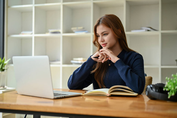 Una joven empresaria asiática inspirada o estudiante universitaria se centra en mirar la pantalla de su computadora portátil, leer un artículo en línea y trabajar en sus tareas en su computadora portátil en una mesa. - Foto, Imagen