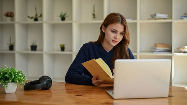Una hermosa joven asiática se centra en leer algo en la pantalla de su computadora portátil mientras trabaja en su tarea en una mesa en un espacio de co-trabajo. - Foto, imagen