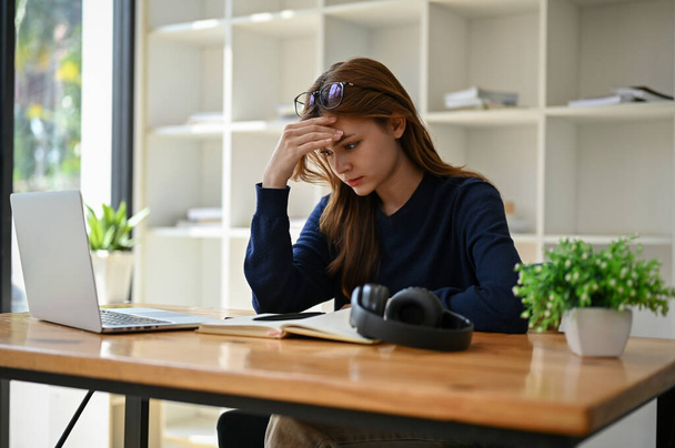Stresowana i przemyślana młoda azjatycka bizneswoman siedzi przy biurku w biurze, próbując znaleźć rozwiązanie swoich problemów w pracy, cierpiąc na ciężką pracę i bóle głowy.. - Zdjęcie, obraz