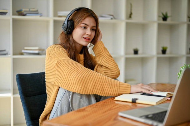 Μια χαλαρή και δροσερή νεαρή Ασιάτισσα γυναίκα σε ένα άνετο πουλόβερ ακούει μουσική μέσω των ακουστικών της, ενώ κάνει την εργασία σε ένα τραπέζι στο σαλόνι. - Φωτογραφία, εικόνα