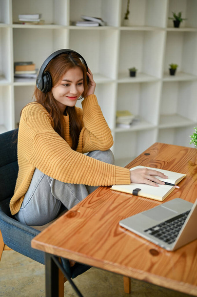 A happy young Asian woman in casual clothes is listening to music through her headphones while doing homework at a table in her room. Lifestyle concept - Photo, Image