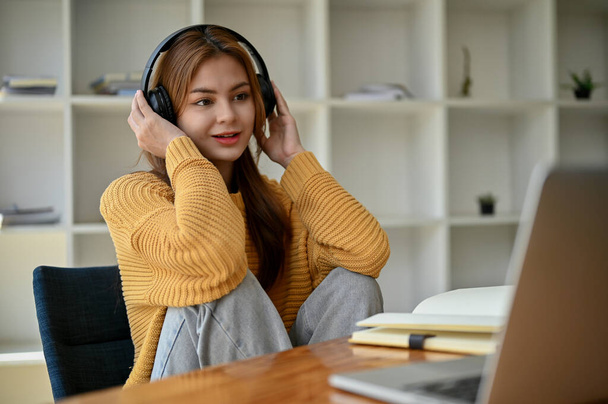 Uma jovem mulher asiática atraente em uma camisola amarela acolhedora ouve música ou podcasts através de seus fones de ouvido enquanto se senta em sua mesa de trabalho em casa. - Foto, Imagem