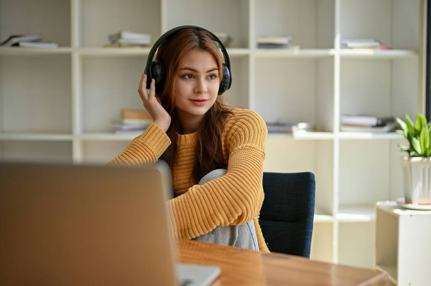 Egy nyugodt és gyönyörű fiatal ázsiai nő egy kényelmes pulóverben zenét hallgat a fejhallgatóján keresztül, álmodozik, és kinéz az ablakon, miközben egy asztalnál ül a kávézóban.. - Fotó, kép