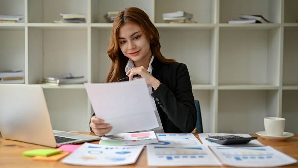 Een aantrekkelijke en lachende jonge Aziatische zakenvrouw of vrouwelijke financiële analist leest een financieel rapport, het bekijken en analyseren van de financiële gegevens op het rapport aan haar bureau. - Foto, afbeelding