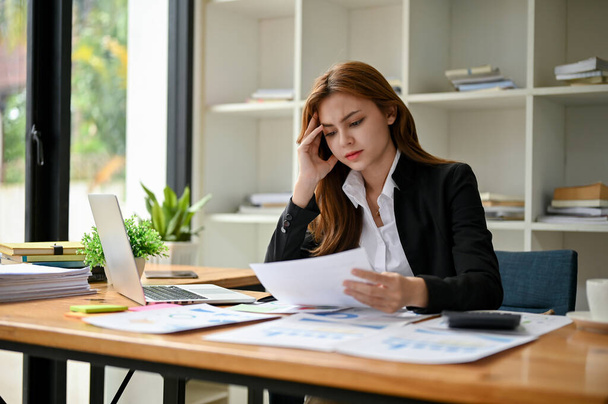ストレスを感じて疲れたアジアのビジネスマンは、彼女のオフィスの彼女の机で財務報告書を読み、分析することに焦点を当てています. - 写真・画像