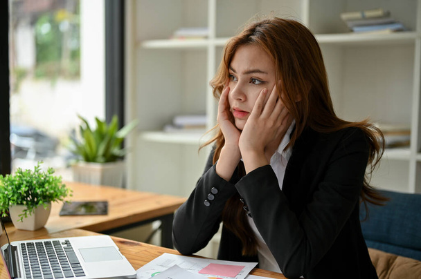 Zestresowana i znudzona młoda azjatycka bizneswoman siedzi przy biurku z rękami na podbródku, wyglądając przez okno z poważną twarzą. zmęczony, wyczerpany, przepracowany, brak pasji w życiu - Zdjęcie, obraz