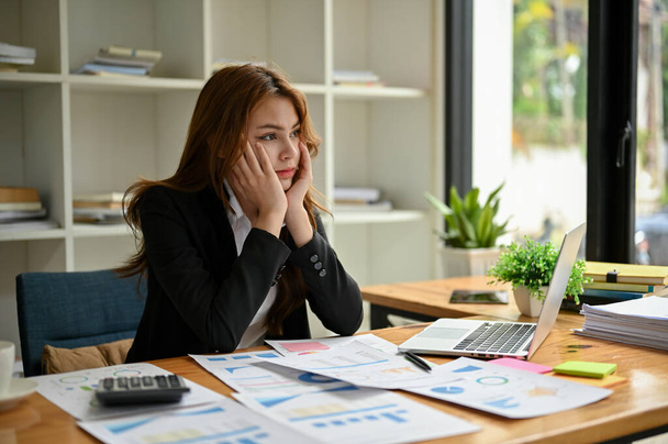 Разочарованная и скучающая молодая азиатская бизнесвумен или финансовая работница сидит за своим столом с руками на подбородке, глядя в окно с серьезным лицом. - Фото, изображение