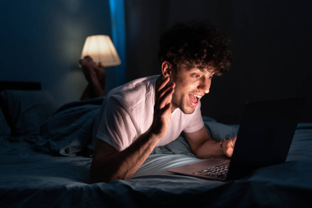 Joven hombre durmiendo y el uso de ordenador portátil para la red social o videollamada en la cama por la noche, estilo de vida y relajarse, el trabajo desde el concepto de casa - Foto, Imagen