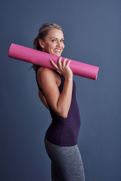 Ready to go to work. Studio portrait of an attractive mature woman in sportswear holding an exercise mat against a blue background - Foto, Bild