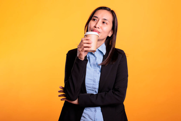Positive asian woman enjoying hot cappucino in studio posing over yellow background, holding cup of coffee during work time. Smiling confident young adult in formal suit serving refreshment beverage - Zdjęcie, obraz