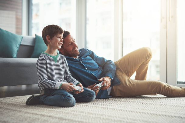 Nunca me vou cansar disto. um menino alegre e seu pai jogando videogames juntos na televisão enquanto estavam sentados no chão em casa durante o dia - Foto, Imagem