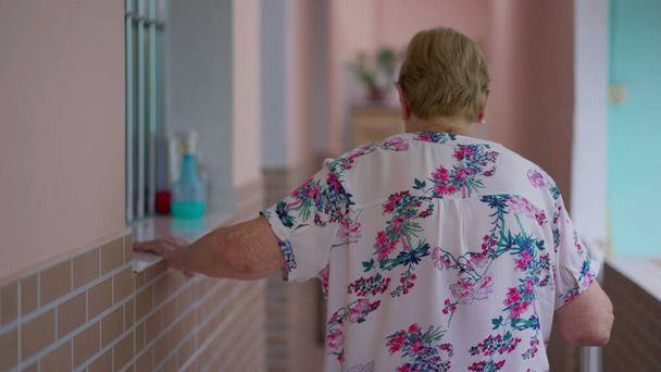 Задняя часть пожилой женщины 80-ти лет, идущей по домашнему коридору, держа стену. Пенсионер на сцене бытового образа жизни - Фото, изображение