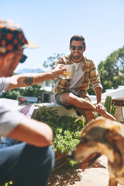Heres to us. two cheerful young men hanging out outside on their balcony with their dog at home - Foto, Imagen