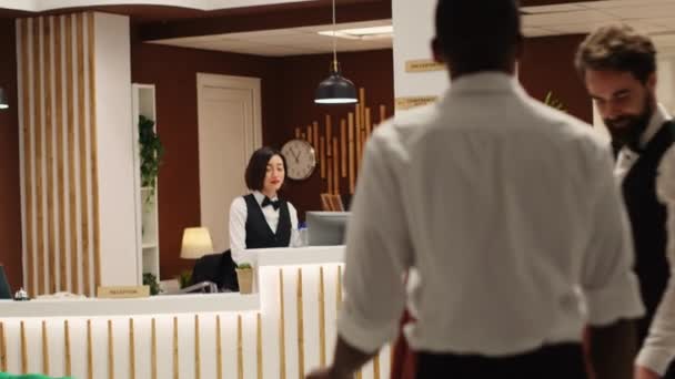 Friendly smiling concierge staff team welcoming guests in hotel lobby. Professional bellboy helping arriving tourists with their baggage while cheerful receptionist provides check in assistance - Footage, Video