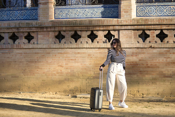 Young and beautiful woman, Hispanic, brown hair, with striped sweater, linen pants and sunglasses, standing, waiting by her suitcase at sunset. Concept beauty, fashion, travel, vacation, luggage. - Foto, Bild