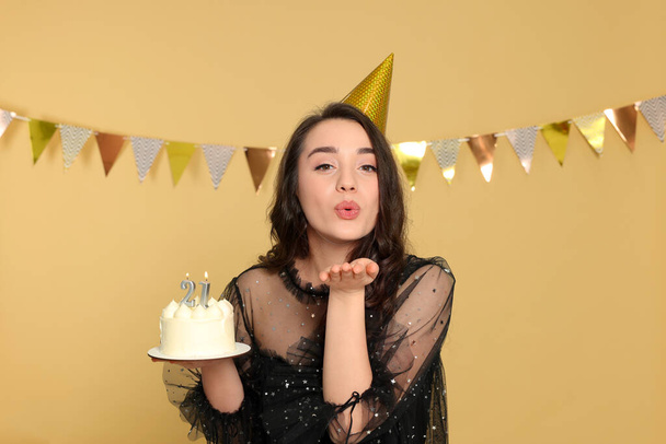 Coming of age party - 21st birthday. Woman holding delicious cake with number shaped candles and sending air kiss on beige background - Fotó, kép
