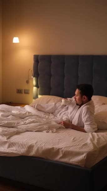 mladý muž v bílém županu pít kávu ráno na posteli - Záběry, video