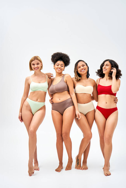 Full length of joyful multiethnic women in colorful bras and panties hugging while standing together on grey background, different body types and self-acceptance concept, multicultural models - 写真・画像