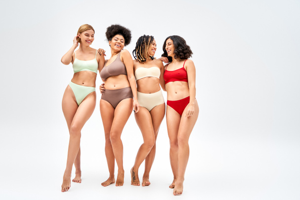 Pleine longueur de heureux groupe multiethnique de femmes en lingerie colorée étreignant et debout sur fond gris, différents types de corps et concept d'acceptation de soi, modèles multiculturels - Photo, image