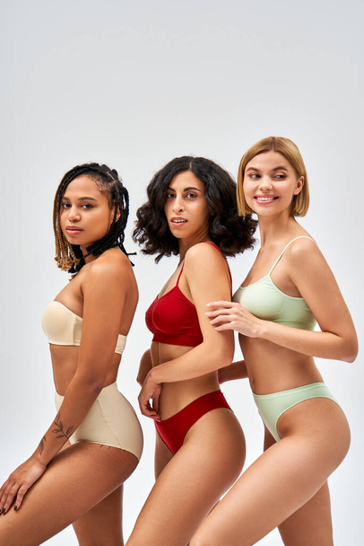 Smiling blonde woman in lingerie looking away while standing near multiethnic friends and posing isolated on grey, different body types and self-acceptance concept, multicultural models - Foto, Imagem