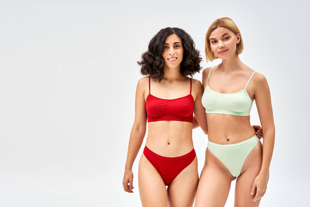 Smiling multiracial woman in lingerie hugging blonde friend and looking at camera while standing isolated on grey, different body types and self-acceptance concept, multicultural models - Photo, Image